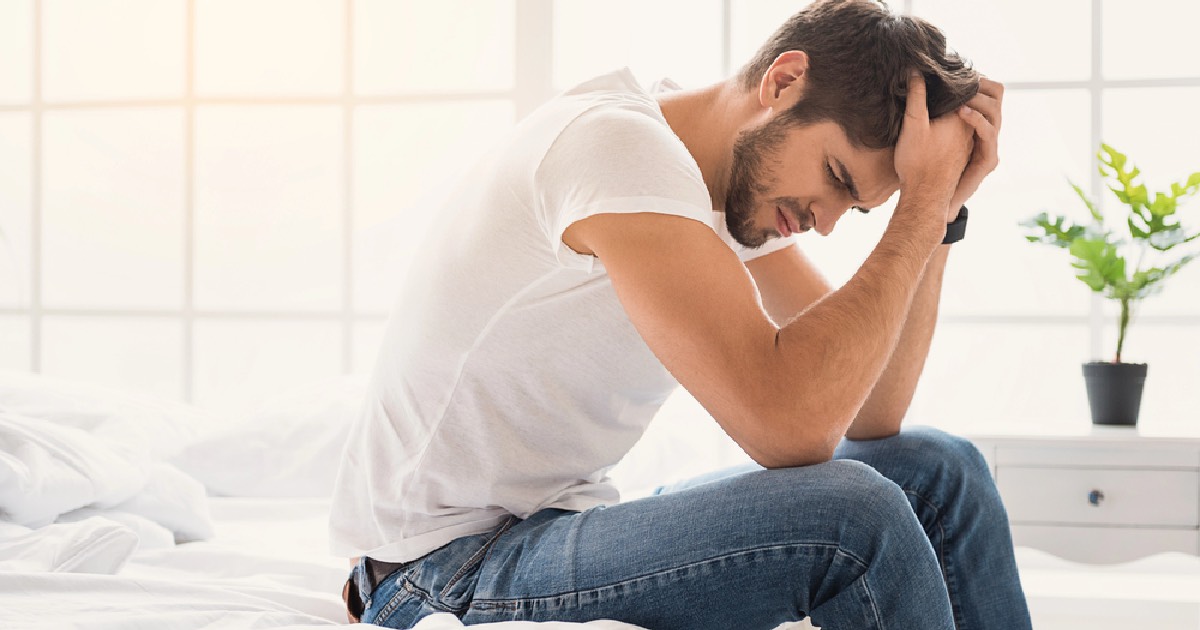 Young man sitting on bed holding his head